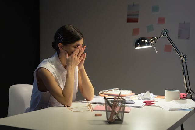 A woman sits at a desk in a dimly lit room, covering her face with her hands, appearing stressed. Papers, notebooks, a lamp, and crumpled paper are scattered on the desk.
