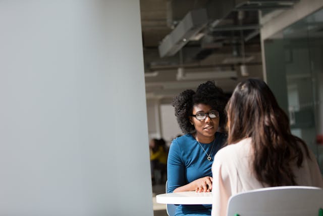 Two women sit across from each other at a round table in an office setting, engaged in a conversation. One woman faces the camera, wearing glasses and a blue top; the other has her back to the camera.