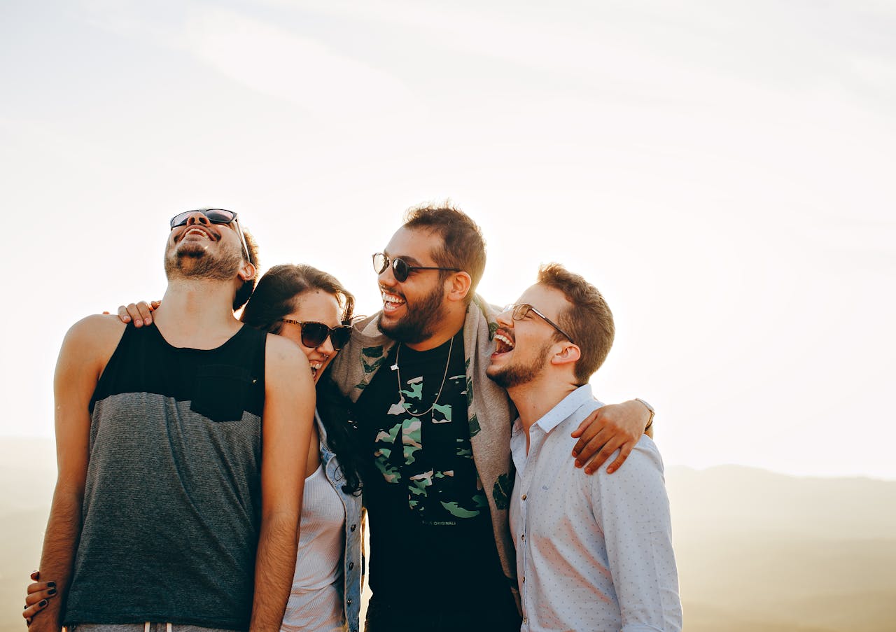 Four friends, two men and two women, stand close together outdoors, smiling and laughing. All are wearing sunglasses, enjoying a sunny day with a bright sky in the background.