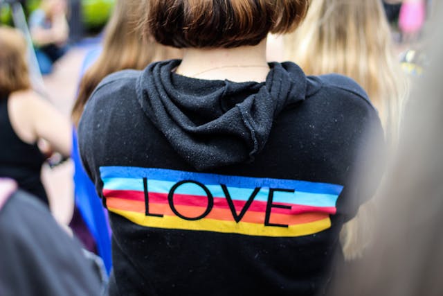 A person with short brown hair wears a black hoodie featuring the word "LOVE" over horizontal blue, pink, and yellow stripes, surrounded by others at an outdoor gathering.