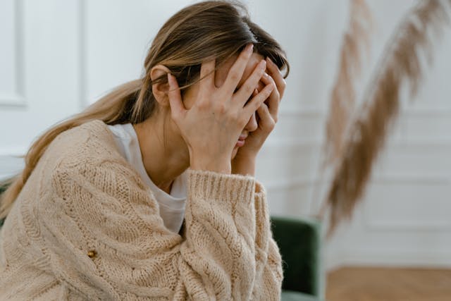 A woman sits indoors with her head in her hands, covering her face, appearing distressed or upset. She wears a beige knitted sweater and has long hair pulled back. The background is softly lit and blurred.
