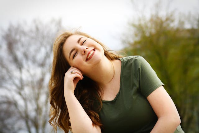 A young woman with long, wavy brown hair smiles with her eyes closed, tilting her head and resting it on her hand. She is wearing a green shirt and is outdoors, with blurred trees in the background.