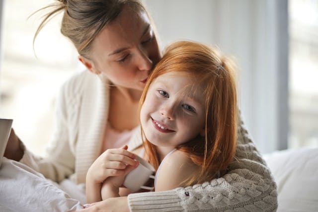 A woman hugs and kisses a smiling young girl with red hair. They are sitting close together indoors, both holding white mugs, with soft natural light coming through a window behind them.
