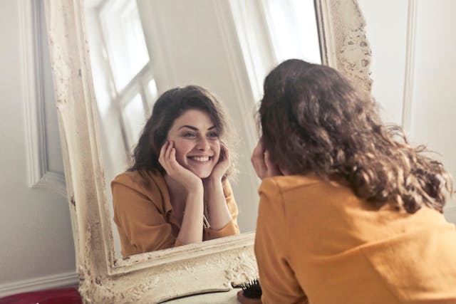 A woman with wavy hair, wearing a yellow top, smiles brightly at her reflection in a large, ornate mirror while resting her face in her hands.
