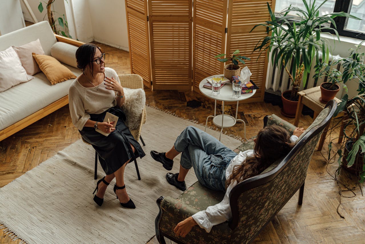 Two women sit in a cozy living room filled with plants and natural light. One woman sits on a chair holding a notebook, while the other lounges on a vintage sofa. A small table with books and drinks stands beside them.