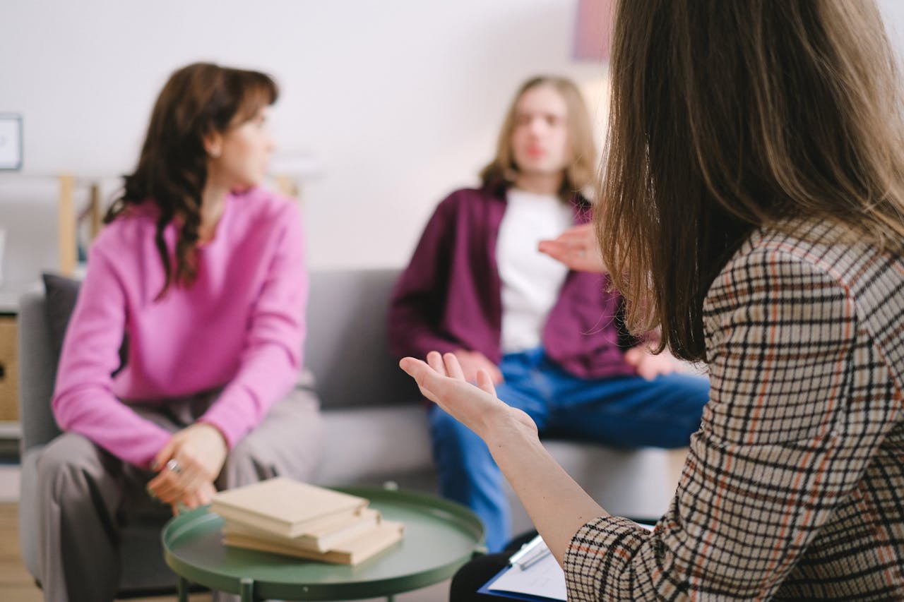 Three people are having a conversation in a living room. Two sit on a couch, one in pink and one in purple, while a third person with long hair gestures in the foreground, facing the group. Books are on a round table.